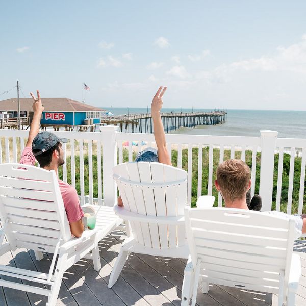 People in white beach chairs overlooking the Atlantic Ocean and the Avon Pier at the Beach Klub at Koru Village's upper deck.
