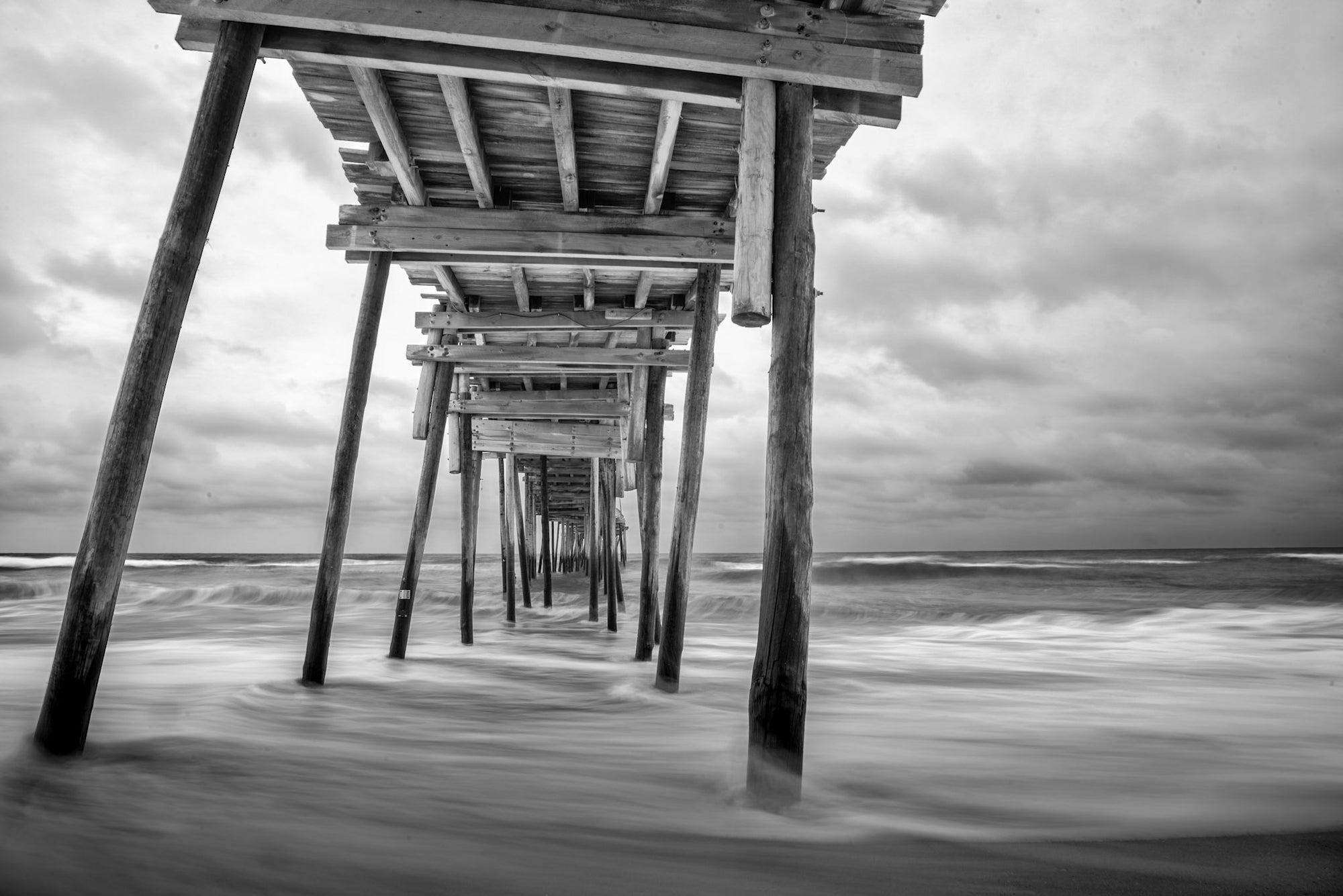 The Avon Fishing Pier in black and white with the waves of the Atlantic Ocean breaking on the Avon beach on the Outer Banks. 